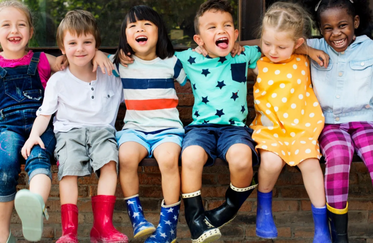 A candid shot of six children of different ethnicities, all smiling and embracing each other while looking in different directions. The children are dressed in bright, colorful clothing, showcasing the diversity of their backgrounds. Their expressions are joyful, highlighting the bonds of friendship and the strength of their social skills, such as empathy, cooperation, and communication. This moment captures the essence of social development in early childhood, where play and group interactions—like those encouraged by Montessori toys—help children build strong relationships and learn to collaborate with others in a supportive, inclusive environment.