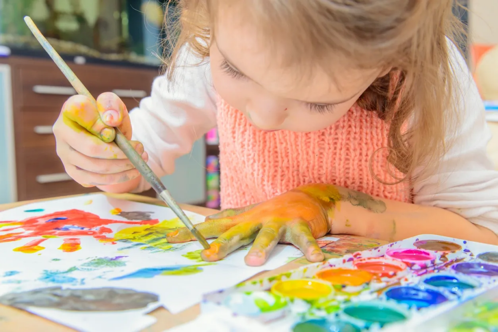 A 3-4-year-old girl is fully immersed in sensory play as she uses a bright acrylic paint set to trace the outline of her hands with a fine-tipped paintbrush. Her hands are covered in vibrant colors—red, blue, yellow, and green—as she explores both the texture of the paint and the shapes of her own hands. The child is deeply engaged in fine motor skill development, using precise hand movements to paint and trace. This activity not only encourages creative exploration but also helps her understand color recognition and body awareness. The hands-on nature of this sensory experience ties directly to the Montessori approach, where tactile engagement and visual stimulation play a crucial role in developing motor skills and cognitive growth in early childhood.