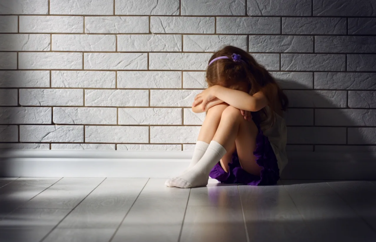 A small girl, approximately 4-5 years old, sits against a white subway tile wall, wearing a purple headband and matching purple skirt. She has her arms folded and resting on her knees, with her head buried in her arms, indicating that she is feeling upset or frustrated. This emotional moment highlights the need for emotional regulation, a key aspect of emotional development in young children. Montessori toys, such as sensory toys or wooden puzzles, can help children like her manage their emotions by providing a calming, hands-on activity to encourage self-soothing and patience. These toys foster emotional regulation and resilience through play, helping children navigate their feelings in a healthy and constructive way.