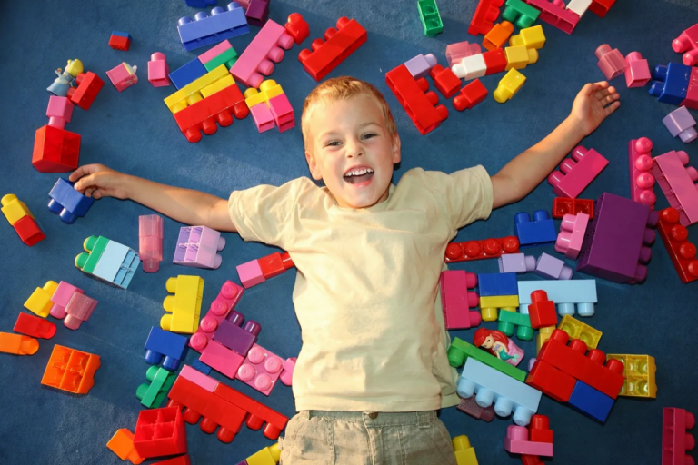 A young boy, around 4-5 years old, lies on a bright blue background, surrounded by colorful manipulatives, including large, easy-to-grip Lego-style blocks. The boy is engaged in fine motor skill development as he picks up and arranges the manipulatives. His focused expression reflects his concentration while using these large, colorful blocks to enhance his hand-eye coordination and dexterity. This Montessori-inspired activity promotes both cognitive growth and fine motor skills, making it ideal for older toddlers learning through hands-on play with large building blocks.