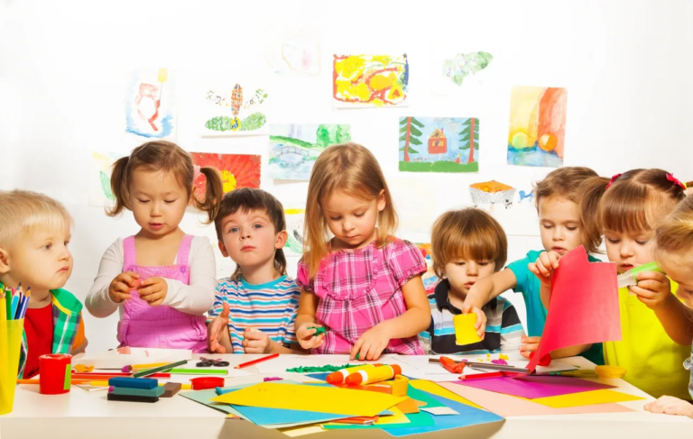 A group of eight children, ages 2-4, are gathered around a vibrant, colorful arts and crafts table, fully engaged in various creative activities. The table is filled with craft supplies like glue, crayons, glitter, play dough, and colorful construction paper. Some children are cutting shapes with safety scissors, while others are using glue to attach pieces of paper or adding glitter to their creations. The atmosphere is lively, with each child working on their own unique project, showcasing their creativity and imagination. This hands-on Montessori-inspired activity encourages independent thinking, fine motor skill development, and self-expression, as the children explore different textures, colors, and materials through open-ended play.