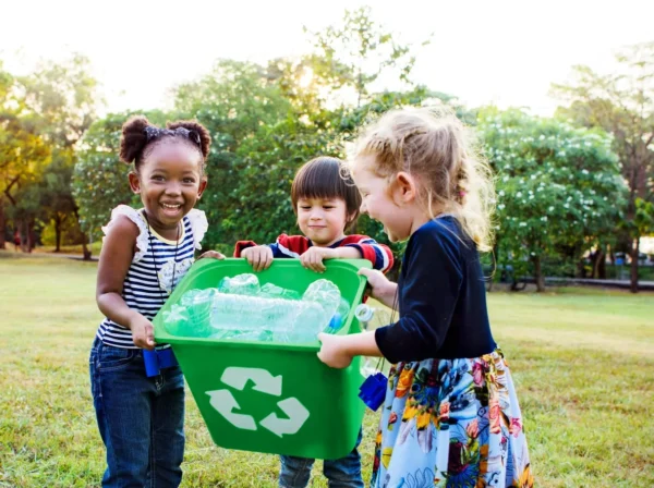 Three children of diverse backgrounds smiling as they collaboratively carry a green recycling bucket, demonstrating teamwork and cooperation. This moment highlights how Montessori toys promote social skills like communication, teamwork, and responsibility, helping children develop essential social skills through cooperative play